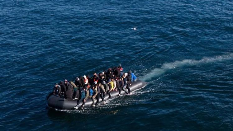 Migrants wearing coats and jackets seen packed onto a black inflatable boat on the English Channel