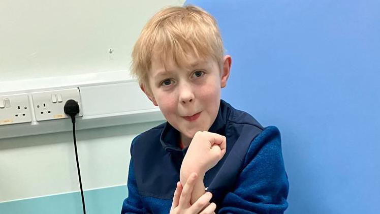 A boy with blonde hair and a blue jumper in a blue hospital chair with a white wall behind him and a plug in a socket.  The boy is holding up one hand to his face.