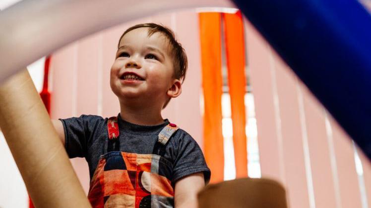 A photo of a boy in a dark grey t-shirt and a multi-coloured pinafore. The boy has brown hair and it smiling, looking above the camera. There is blue and brown play equipment in front of him, blurred by the camera. There are orange blinds behind him in the background. 