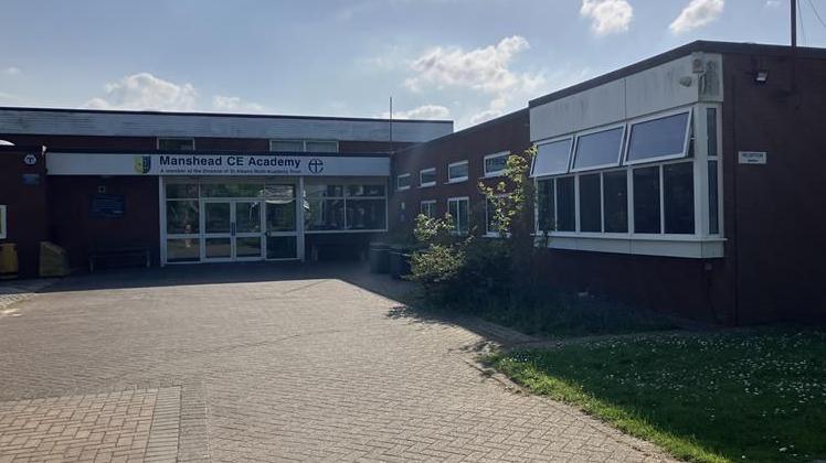 A red bricked building goes across the image. On the left there are two white doors which has a sign above that says Manshead CE Academy. On the right is a large white window and grass out the front of the building. 