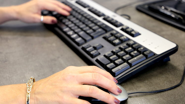 A woman holding a computer mouse and hovering her hand over a keyboard on a grey desk.