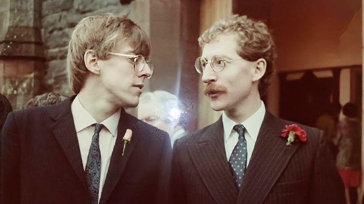 Tow men in suits at a wedding - both men have brown hair and are wearing glasses. They are standing outside a church entrance and looking at each other while in conversation.