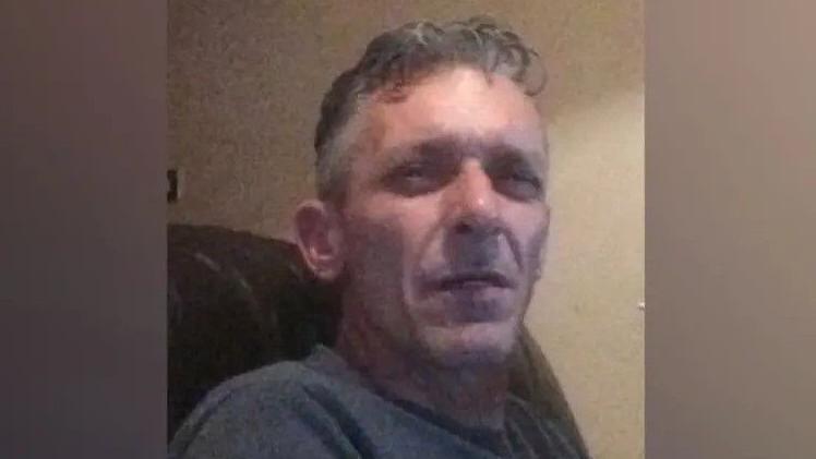 A man in his mid-50s with grey hair. He is sitting in an armchair and squinting at the camera.