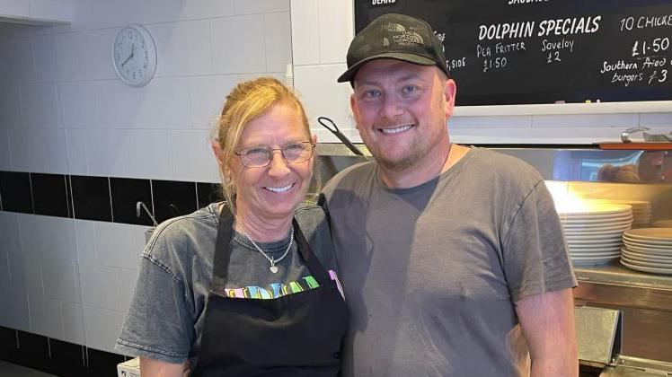 Sharon Farrell, who has blond hair and glasses and is wearing a black apron and grey t-shirt, is side by side with Tony Farrell with one arm round each other's backs. Mr Farrell is wearing a grey t-shirt and black baseball cap. Both are standing in front of the fryers in their fish and chip shop The Dolphin in Fleetwood. 