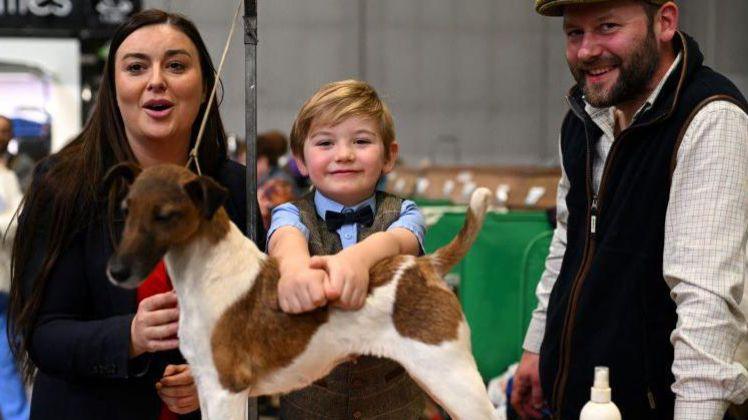 A woman with a black blazer and red dress stands next to a boy in a brown tweed suit and a man in a black waistcoat, white shirt and yellow hat. The boy is holding onto a brown and white fox terrier in front of him.