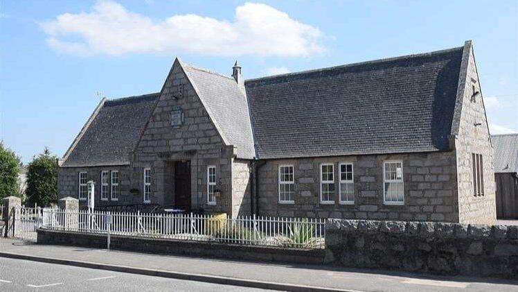 Insch Library - a grey brick building with large sloping roof in front of a white fence. 