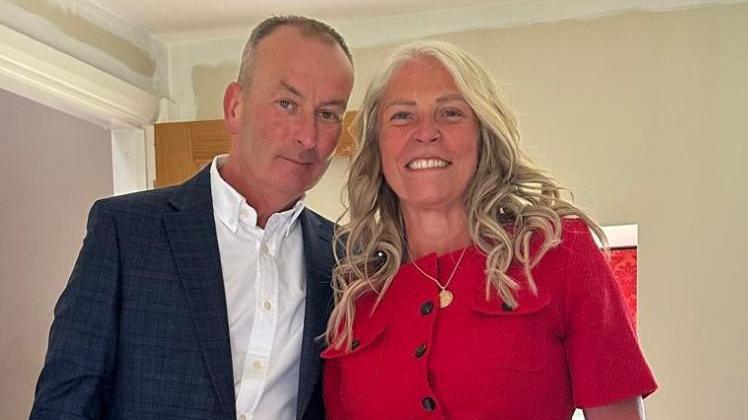 Roger and Lisa stood side by side, looking into the camera and smiling. Roger is wearing a dark blue suit jacket and a white collared shirt. Lisa is wearing a red dress. They're stood indoors in what looks like a living room with beige walls.
