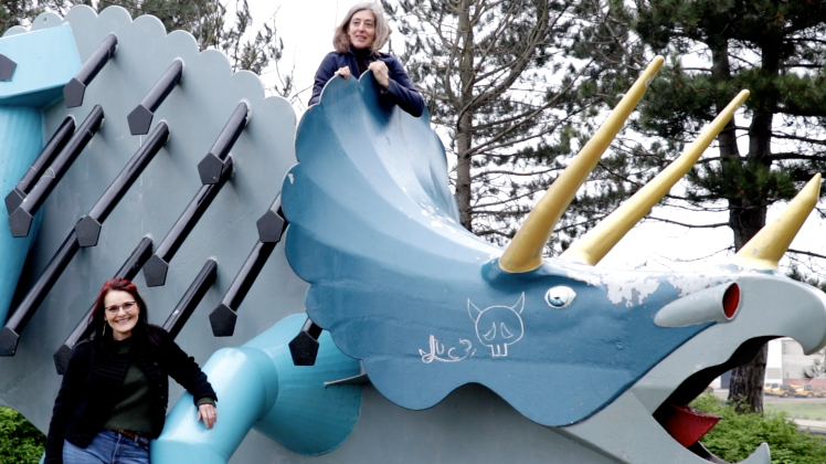 Jude Keenan and Susie Glatt pictured with a large steel triceratops which is painted blue and has graffiti on it. One of the women has climbed to the top of the sculpture.