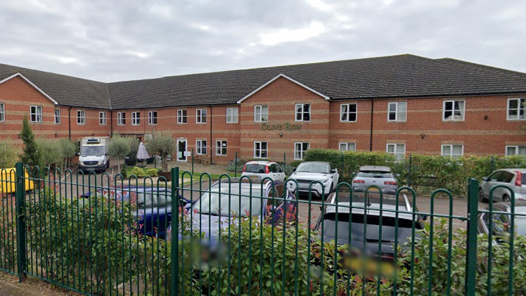The outside of Olive Row, a care home in Kettering. The building is a brown brick residential building with a car park outside of it. The building backs onto a road and has a metal fence and bushes outside of the parameter. 