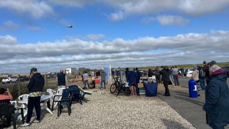 Aviation enthusiasts, some with plastic chairs, standing in a viewing area across the road from RAF Waddington as a plane flies overhead