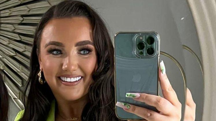 Chloe Ferris smiling at the camera while taking a selfie.  She has long, wavy dark hair and long, painted fingernails.  She wearing make-up, earrings and a lime green outfit. 