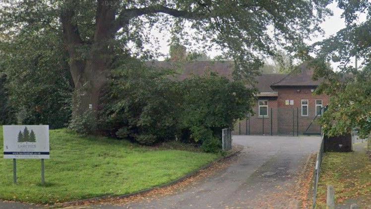 The entrance to Larches High School with gates and a sign saying Larches High School and with an image of three Larch trees. The school building can be seen beyond the gates, surrounded by trees.