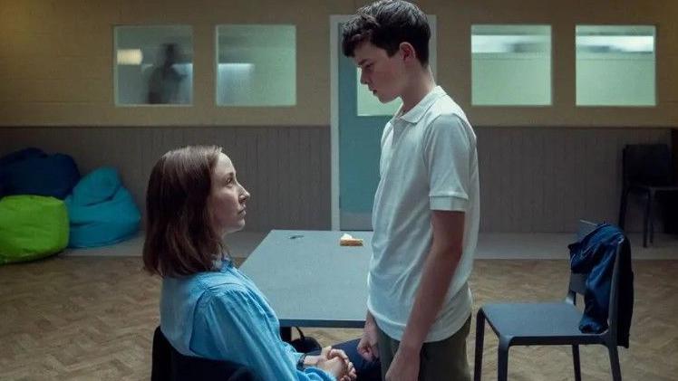 A photograph of a boy in a white shirt standing over a seated woman in a blue blouse. 