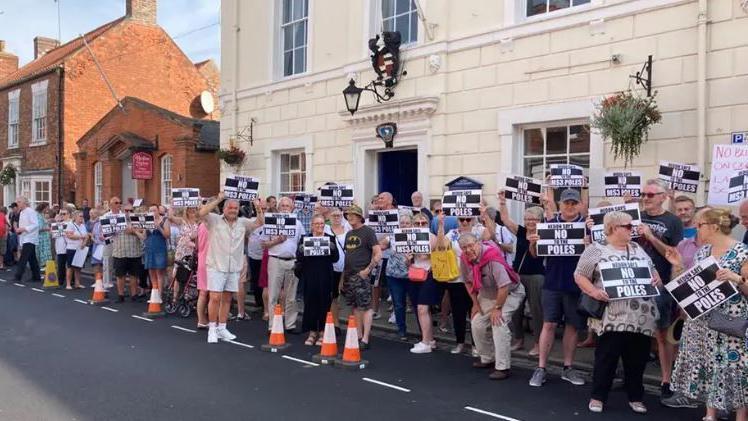 A row of several people at a protest holding up black-and-white signs that say 'no to the poles' on them. They are stood at the side of a road in front of buildings.