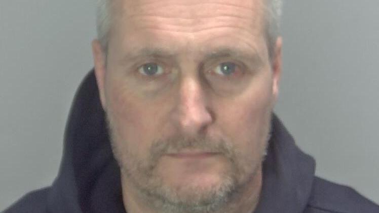 Oliver Arnold has short grey receding hair and a stubbly beard and moustache. He appears on this police custody photo wearing a blue hooded top. The background is grey.
