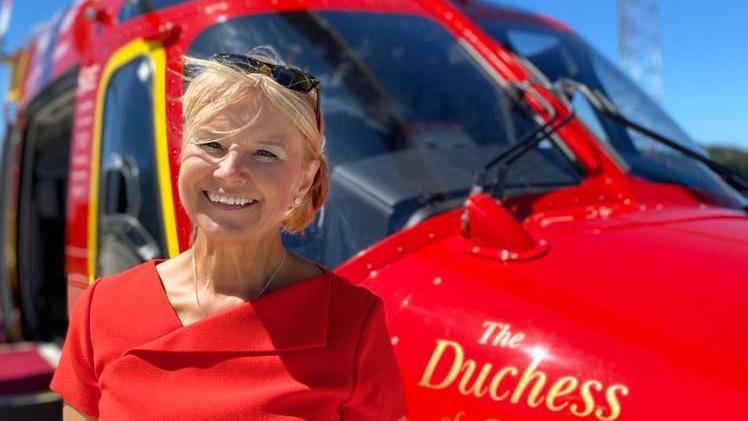 Barbara Sharples wearing a red dress and standing in front of a red Cornwall Air Ambulance. 