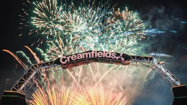 A Creamfields metal sign in the sky on an arch. It is night time and there are huge fireworks behind the black sky going off. 