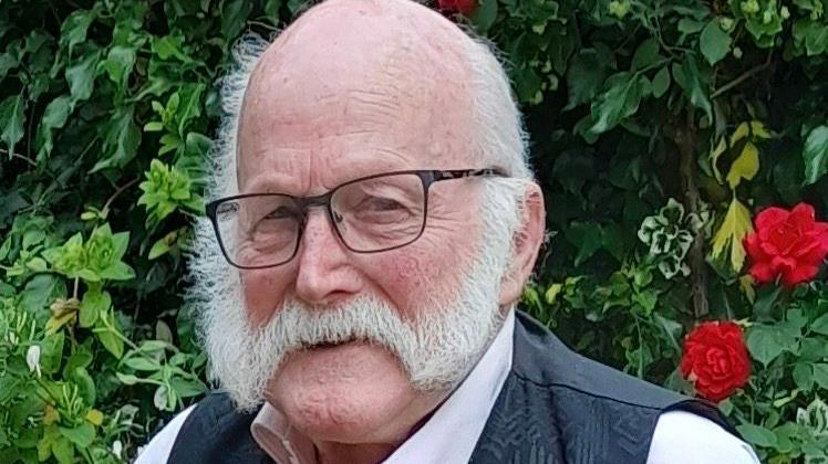 Ray Savage smiles as he stands in front of a bush. He is largely bald with some grey hair on the side of his head and he has a grey beard. He wears glasses with a dark waistcoat and pink shirt.