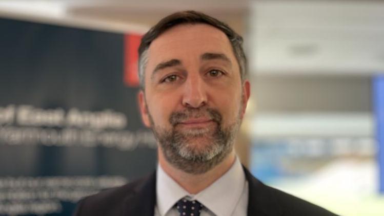 Richard Goffin was in the middle of the image looking towards the camera with a light smile. He had short black and grey hair and beard, and was wearing a white shirt, with a black suit jacket and black and white spotted tie.