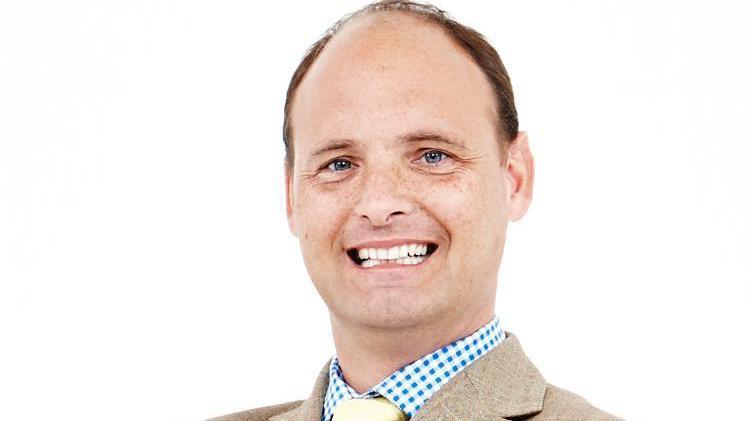 A portrait shot a John Quemard. He has short brown hair and blue eyes and is smiling. The background behind him is white. The picture is of him from the shoulders up. He is wearing a blue and white top and