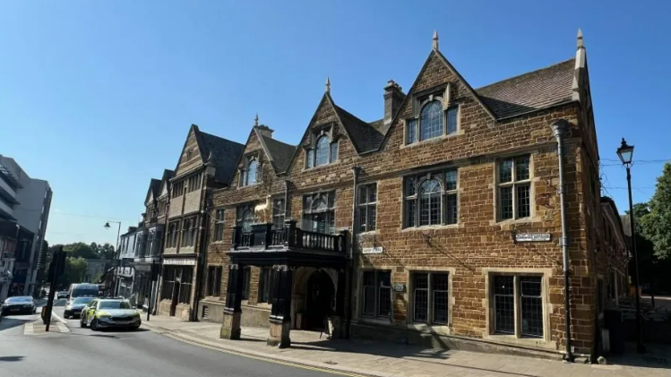 The frontage of the Hind Hotel in Wellingborough - a brown, listed brick building with a wooden canopy outside the main entrance that goes over the pavement. Cars queue at traffic lights on the road outside.