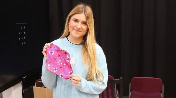 Ella Lambert holding a reusable period pad which is pink with flowers on it. She has blonde hair and is wearing a blue jumper and smiling at the camera