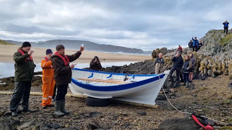 People stand around the boat with glasses raised, toasting its launch.