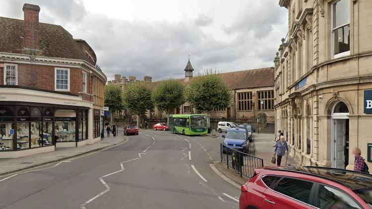 Part of Corportation Street, Taunton, with a red SUV in the corner, red brick and Georgian style building, with the road twisting round into a bend and bus parked up. A cafe on the right and a jewellers shop on the left.