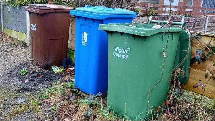A brown, blue and green wheelie bin lined up against a fence with Wigan Council branding on in white lettering