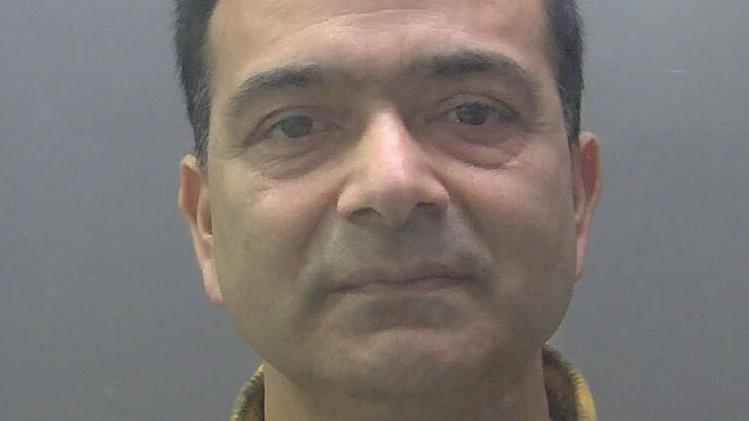 A custody image of Pramod Dhakal. He has short dark hair and is looking into the camera. He has a yellow and black checked shirt collar.