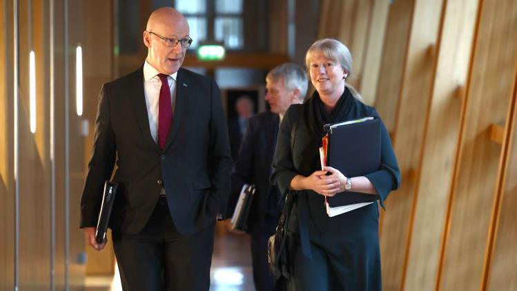 Shona Robison and John Swinney walking down a corridor in the Scottish Parliament. Both are holding black folders under their arms and are dressed in dark suits. Swinney is wearing a red tie and glasses. 