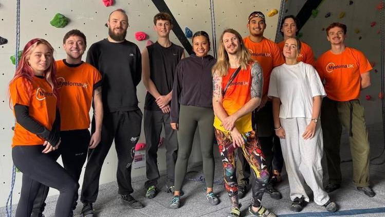 A team of 10 climbers. Some are dressed in orange Myeloma UK t-shirts and standing in front of climbing wall alongside James Moore who has long hair and is wearing brightly coloured trousers.