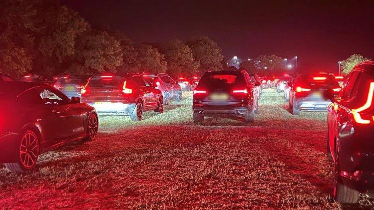 Several long lines of cars queueing up on a field. It is dark. The cars' tail-lights are shining red.