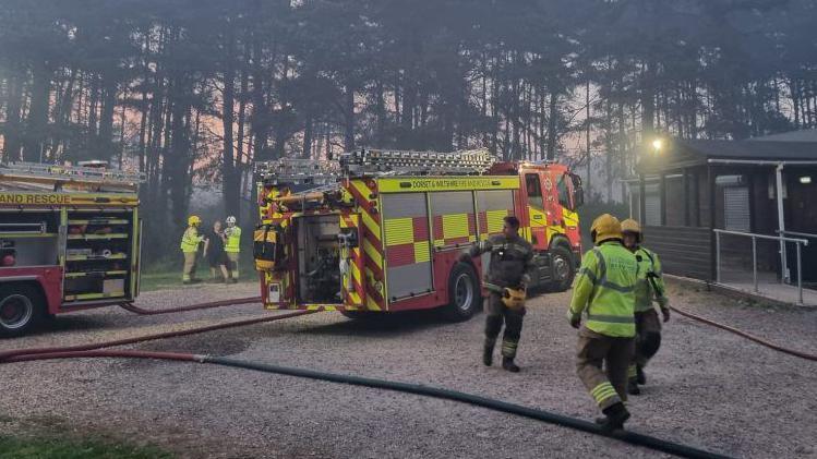 A clearing in a forested area, with a wooden building to the right. There are two fire engines with hose reels leading off camera. Firefighters walk about the scene.