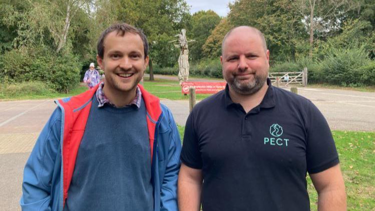 Philip Thomas wearing a blue jacket with a red collar and a blue vest sweater - standing next to Stuart wearing a black T-shirt with a green PECT logo - with green trees and bushes in their background.