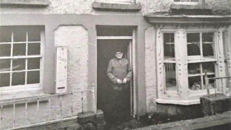 Llun du a Gwyn o hen siop, hen ddyn yn sefyll yn y drws