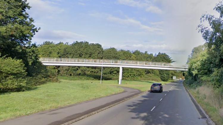 A grey bridge runs over a single carriageway road with a car driving along.