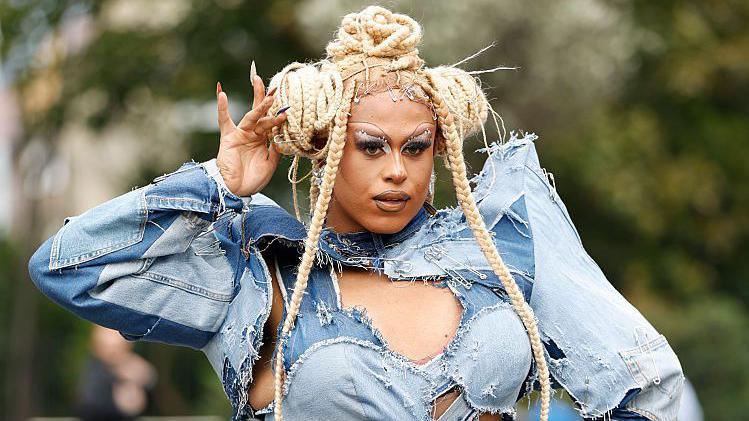 Tayris is posing with her right hand in her hair. She is wearing a denim  strapy top with sleeves. She has a blond wig on which is in plats.