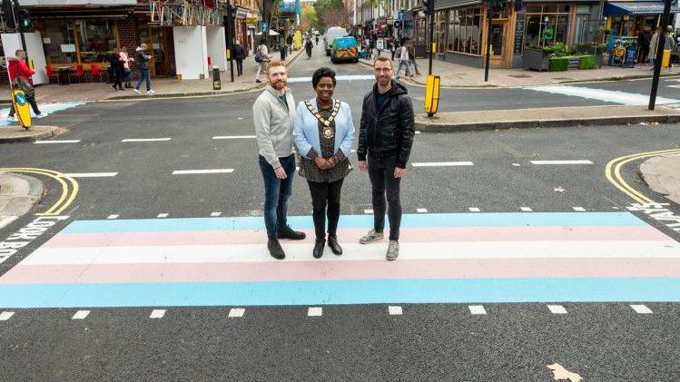 Three people stand on a road crossing in a London street: the crossing is painted in blue, pink and white stripes. They are casually dressed men and a woman who is wearing a blue jacket and a mayoral chain of office. 
