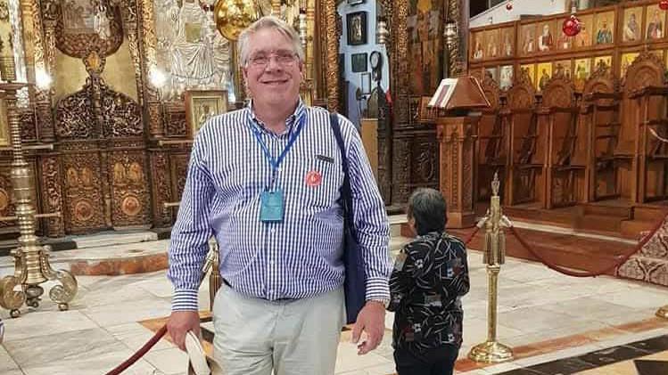 Philip Fryer is wearing a purple checked shirt and beige trousers. He has a lanyard around his neck and is carrying a navy bag on his shoulder