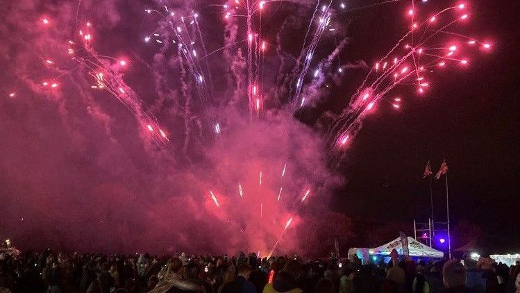 A crowd gathers for a huge fireworks display. Red sparks are visible as well as smoke. A tent in the background is flying two Union Jack flags.