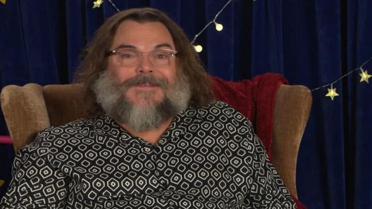 A man with brown hair, glasses and a grey beard is sitting in a brown chair. He's wearing a black and white shirt, and behind him is a blue curtain and fairy lights
