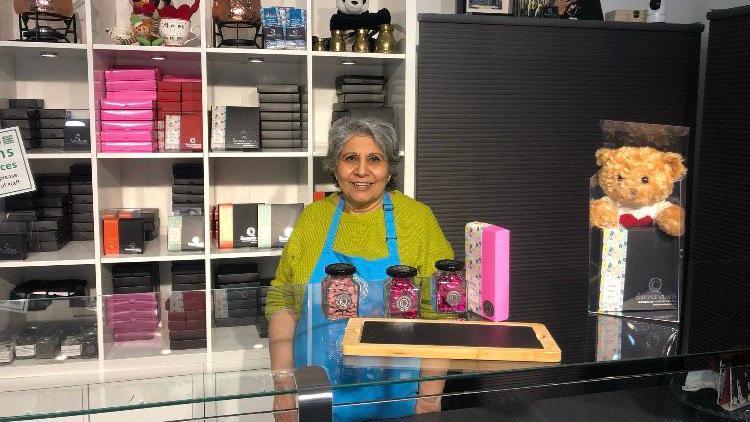 A woman standing behind a chocolate shop counter. She is wearing a green jumper and blue apron. She is smiling at the camera and has thick, grey hair. Boxes of chocolate are in an open, white cupboard behind her. 