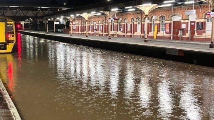 Brown water floods the train tracks at a train station. A yellow train can be seen in the left of the picture. The water has completely submerged the train tracks.