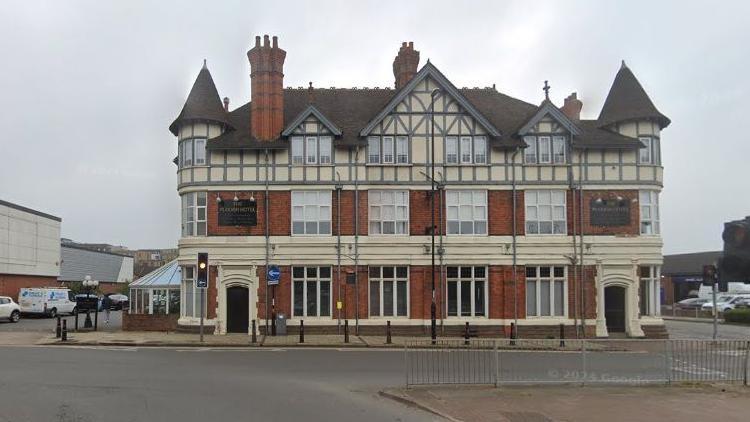 Three-storey brick-built late-Victorian hotel with witch's hat designs on either end of the roof, and three visible chimney stacks. There are windows made up of several small pains evenly spaced across the facade, with taller ones on the ground floor. There are entrance doors on either end of the front facade. An urban road passes in front of the hotel.