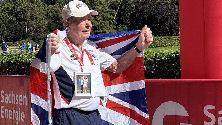 An 89-year-old man wearing Team GB sports uniform and a white cap, holding a union flag behind him, and wearing a photograph of himself and his wife around his neck.