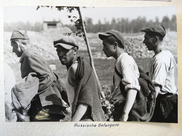 Four men walk in a line, wearing rolled-up shirtsleeves and caps. In the distance, a structure which may be a fortified position can be seen.