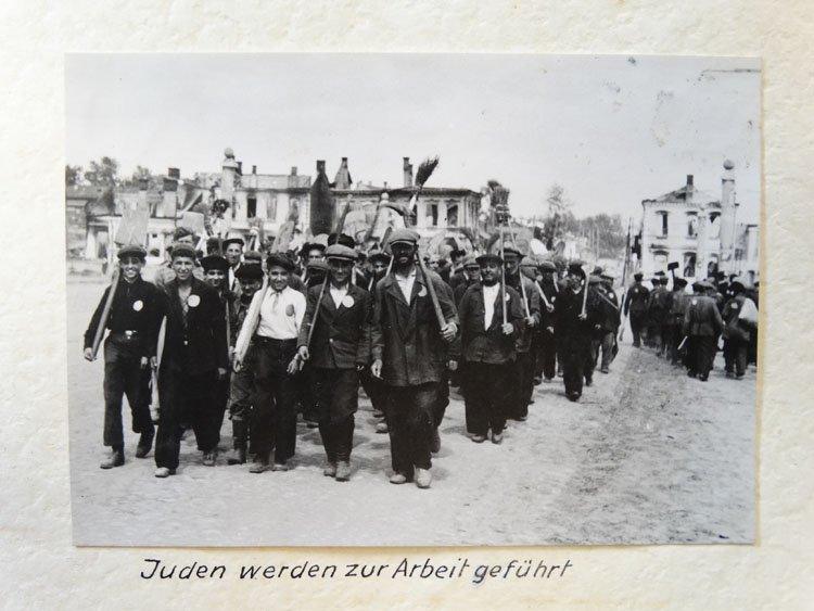 A large crowd of people, wearing the badge marking them as Jewish prisoners, walk in lines, carrying brooms and other work tools.