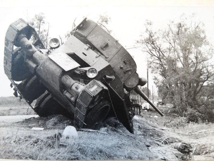 A tank lies toppled at a 45-degree angle on a grassy slope, its door open