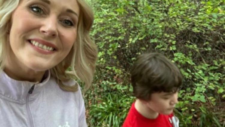 Harry with his mum Deirdre. They are outside walking with green leaves in the background. Harry has brown hair and wears a red top. Deirdre has blonde hair and wears a grey top.